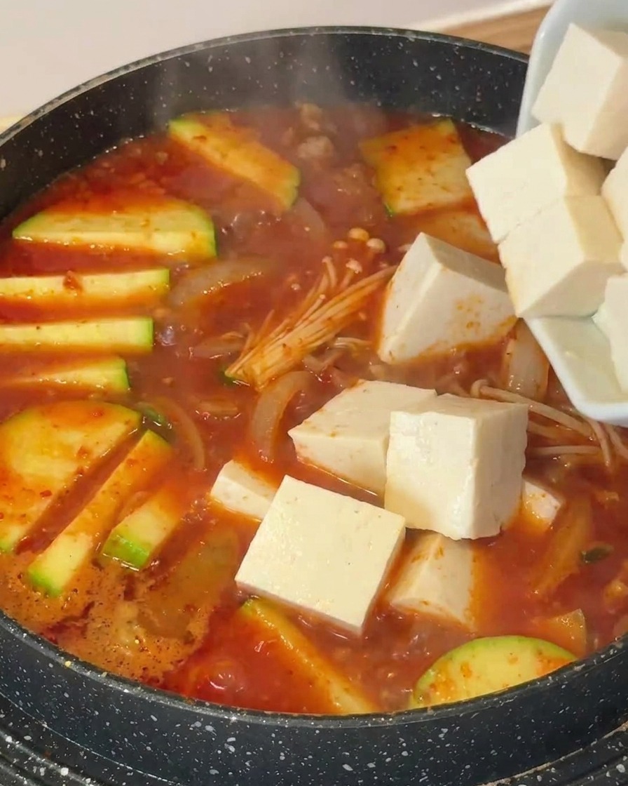 Cubos de tofu blanco añadiéndose a una olla con sopa, calabacín y setas enoki.