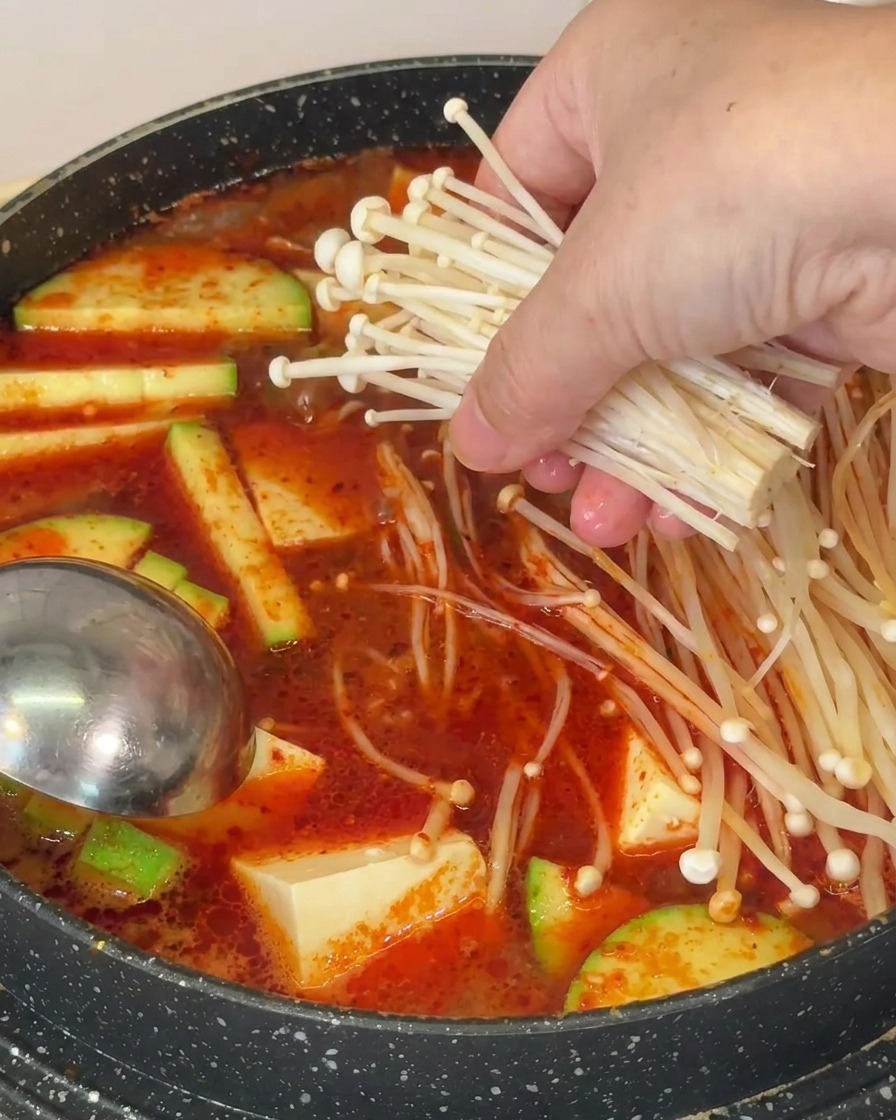 Una mano poniendo un manojo de setas enoki finas en una olla de sopa roja con tofu y calabacín.
