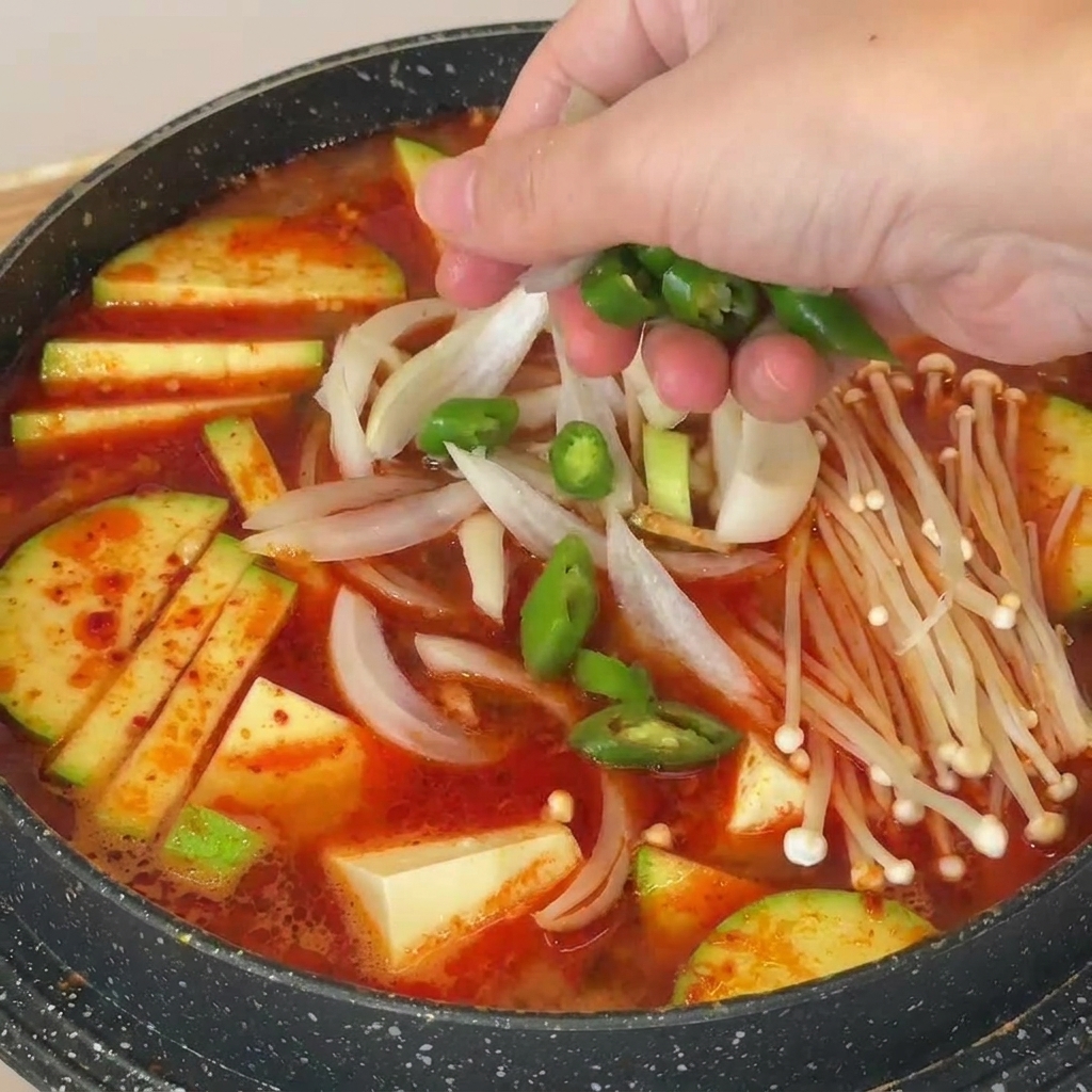 Mano espolvoreando rodajas de chile verde sobre una olla de sopa coreana con setas, tofu y calabacín.