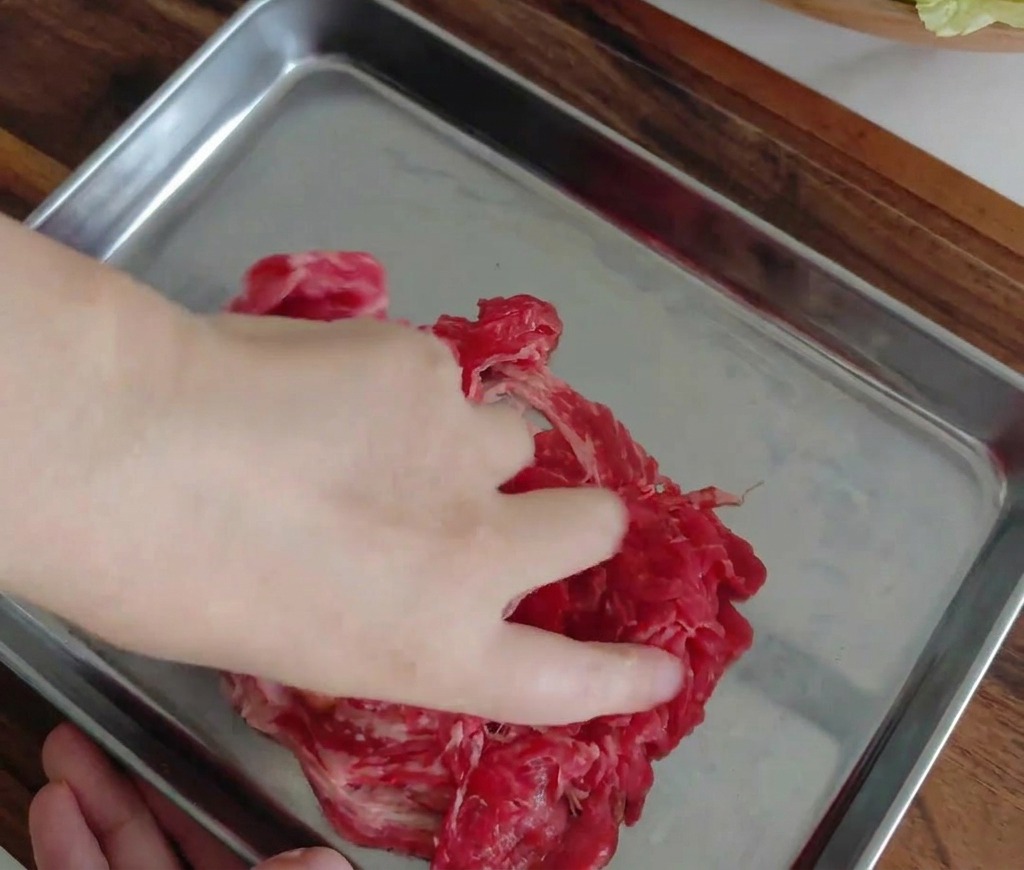 Una mano mezclando las láminas de ternera marinadas en una bandeja de metal.