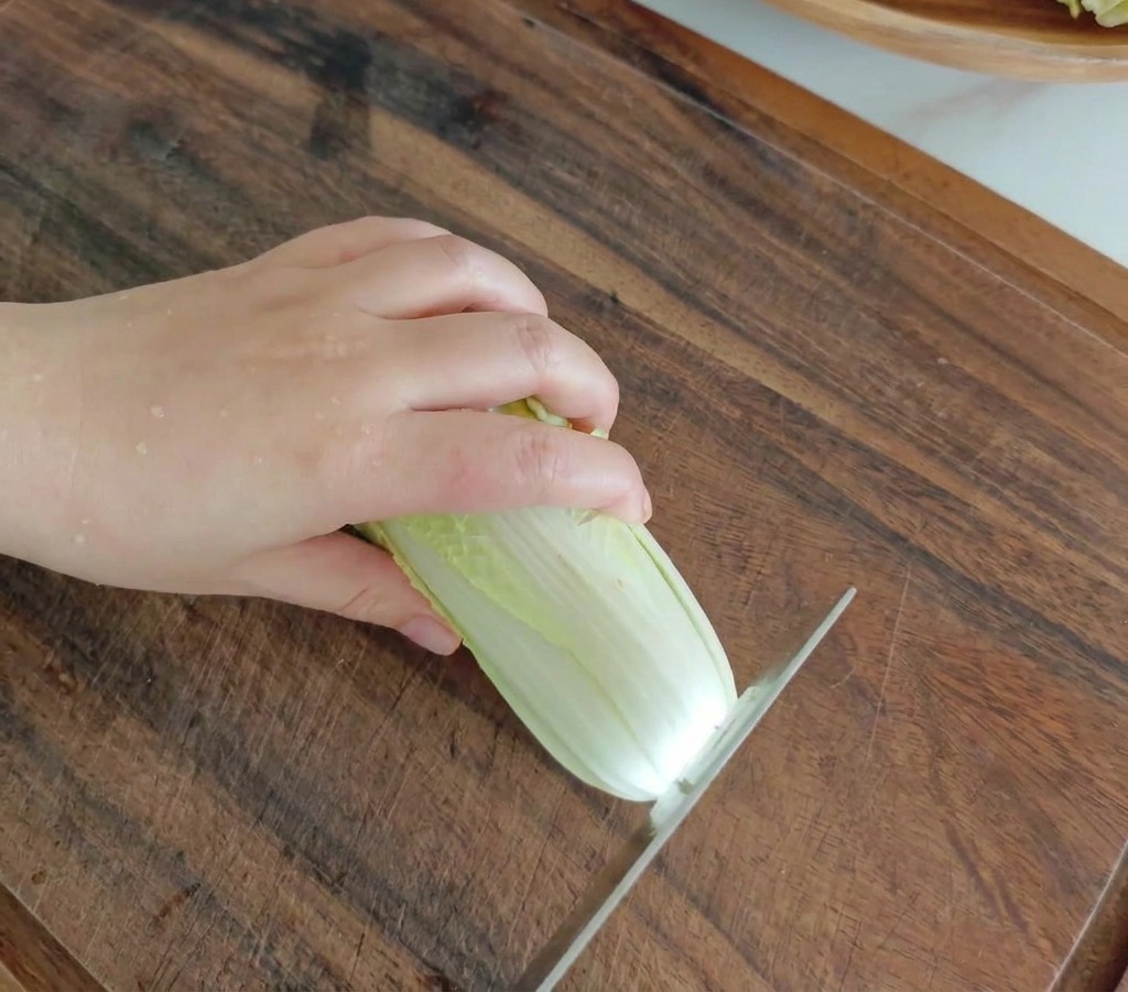 Un cuchillo de cocina cortando una cabeza fresca de col baby sobre una tabla de madera oscura.