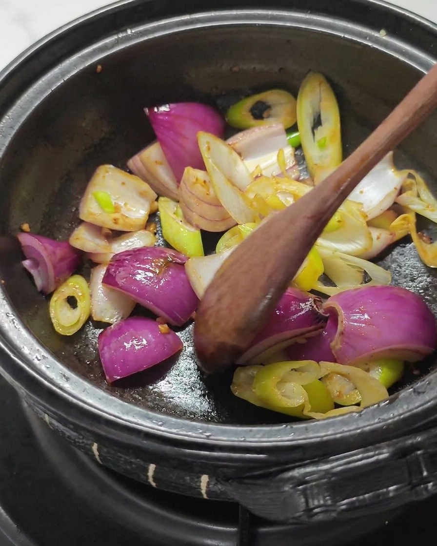 Cuchara de madera removiendo trozos grandes de cebolla morada y blancos de cebolleta en una olla de piedra oscura.