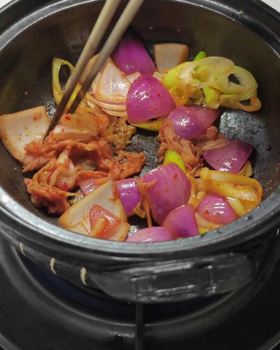 Palillos mezclando kimchi de col picado y pasta de chile rojo con las cebollas salteadas en una olla de piedra.