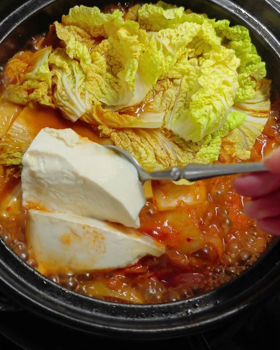 Una cuchara grande bajando suavemente tofu blanco suave en un estofado rojo caliente y burbujeante con hojas de col.
