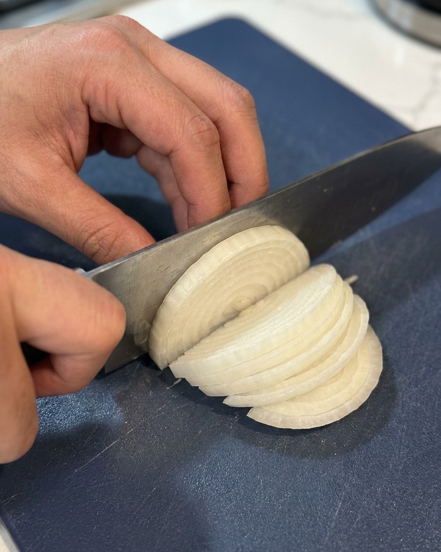Primer plano de manos cortando una cebolla blanca en tiras finas sobre una tabla de cortar azul oscuro.
