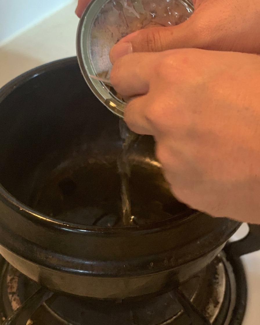 Mano vertiendo el aceite transparente de una lata de atún plateada en una olla de cerámica negra.