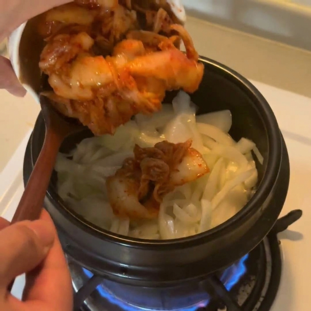 Kimchi de col picado cayendo desde un bol de cerámica blanca hacia la olla con las cebollas salteadas.