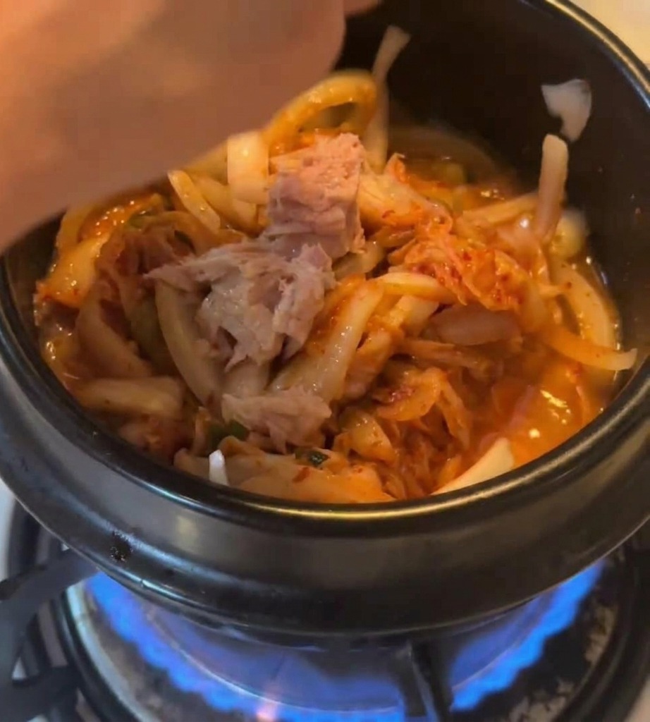Trozos de atún en conserva siendo añadidos a una olla con kimchi y cebollas sobre una llama de gas azul.