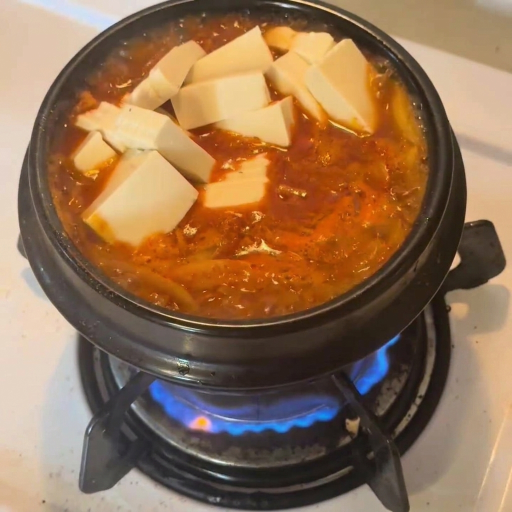 Cubos de tofu blanco fresco reposando sobre el estofado de kimchi rojo en una olla de cerámica.