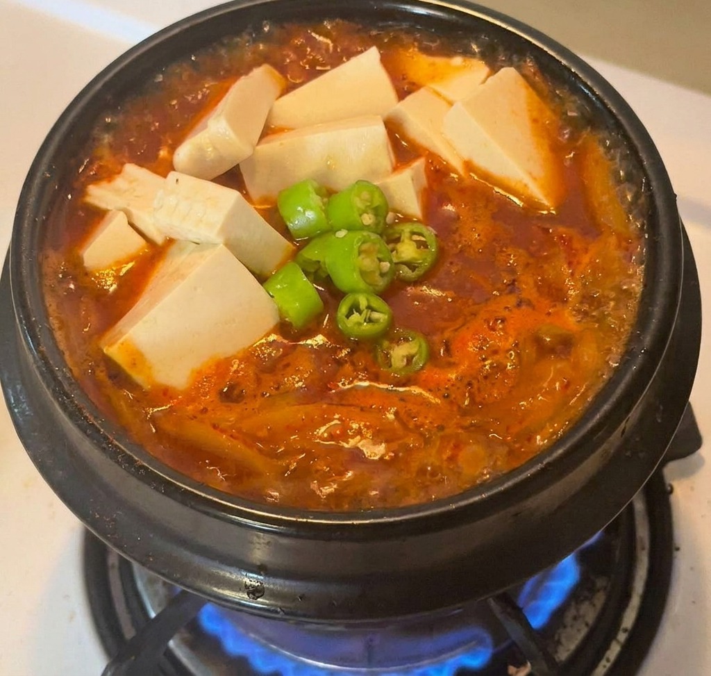 Olla de piedra con estofado rojo, coronada con tofu blanco y rodajas de chiles verdes en el centro.