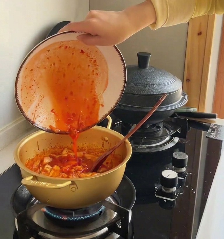 Jugo de kimchi rojo vertiéndose desde un bol de cerámica a la olla con la col salteada.