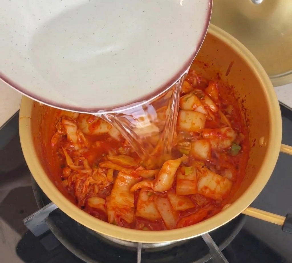 Agua vertiéndose desde un bol a la olla dorada que contiene el kimchi salteado.