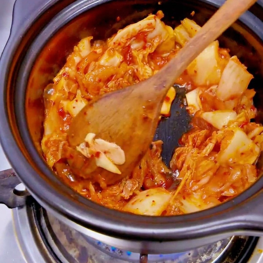 Mano con guante negro dejando caer rodajas de cebolla blanca en una olla con kimchi rojo.