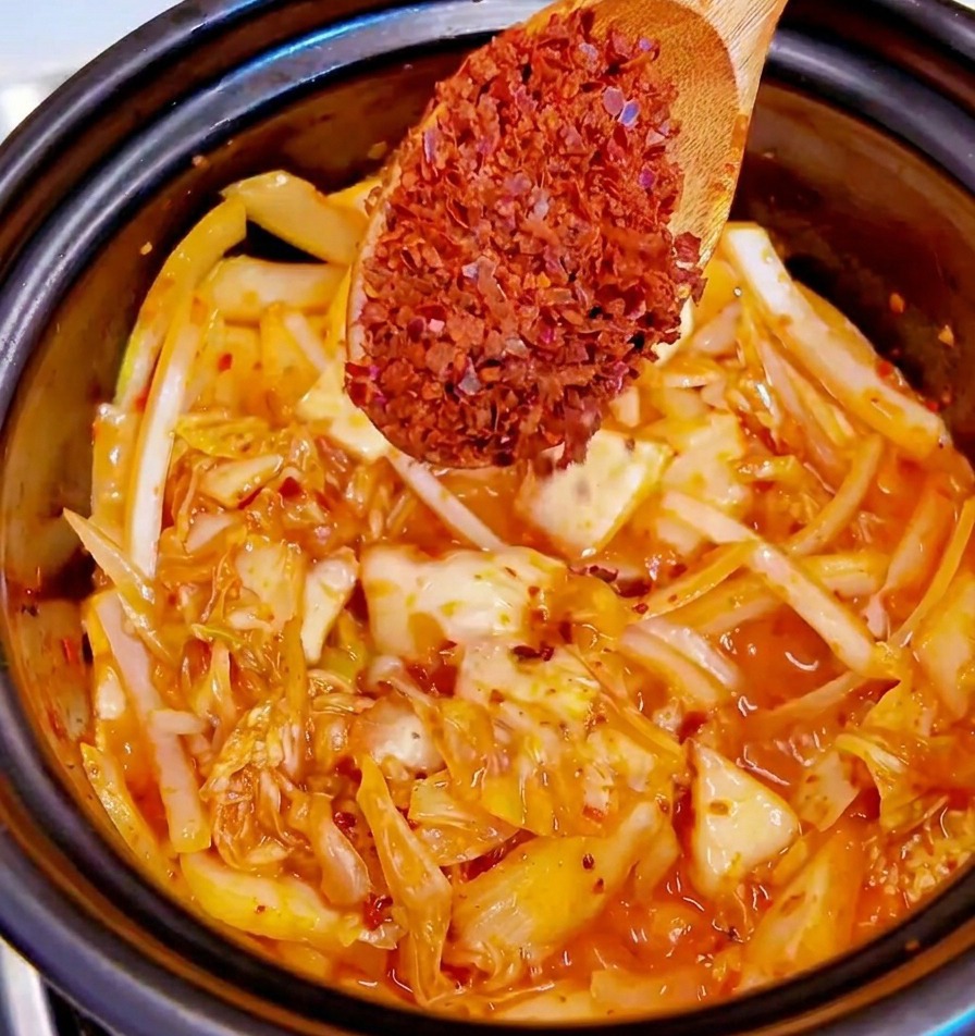 Cuchara de madera con copos de chile rojo sobre una olla de kimchi y cebollas.