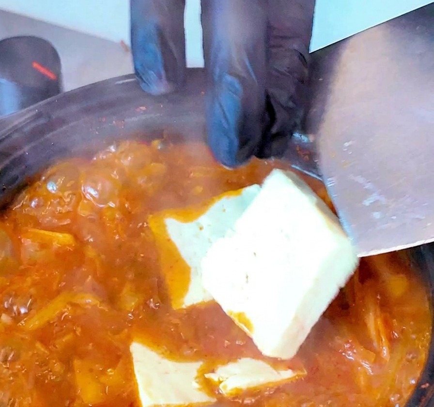Rodajas de tofu blanco introducidas en una sopa de kimchi roja hirviendo con una espátula.