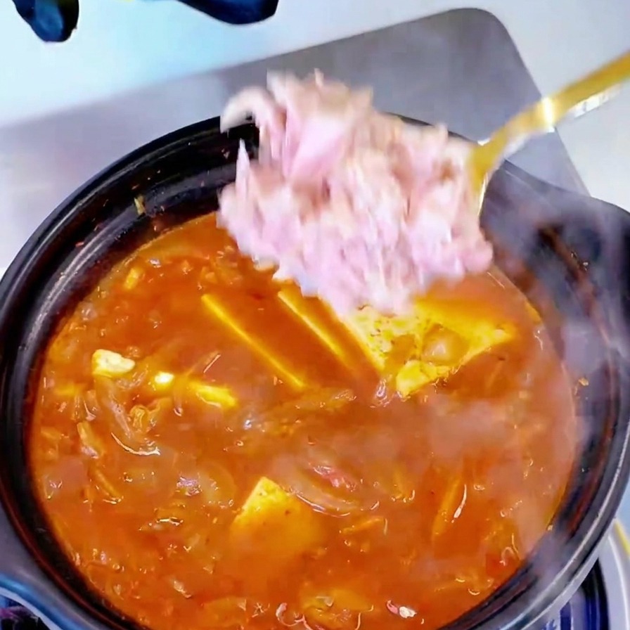 Trozos de atún enlatado añadidos con cuchara a una olla de sopa de kimchi y tofu hirviendo.