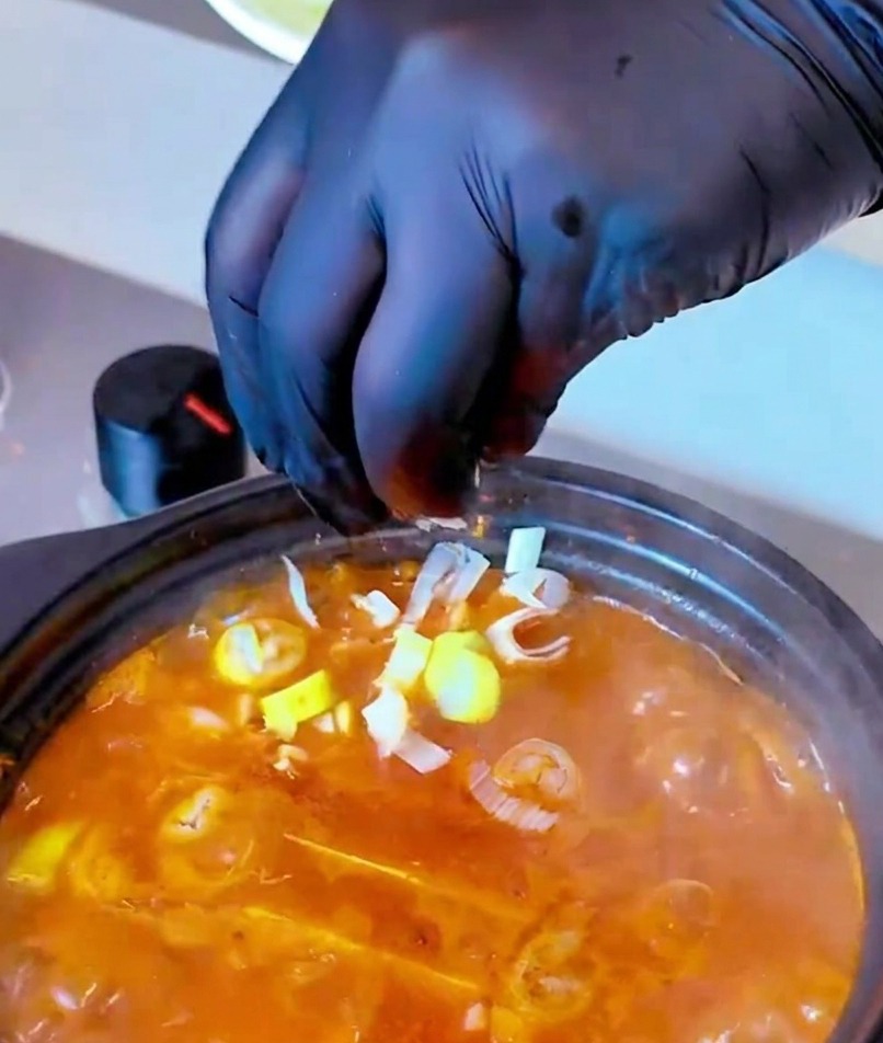Mano con guante negro espolvoreando cebolleta picada fresca en una olla humeante de sopa roja.