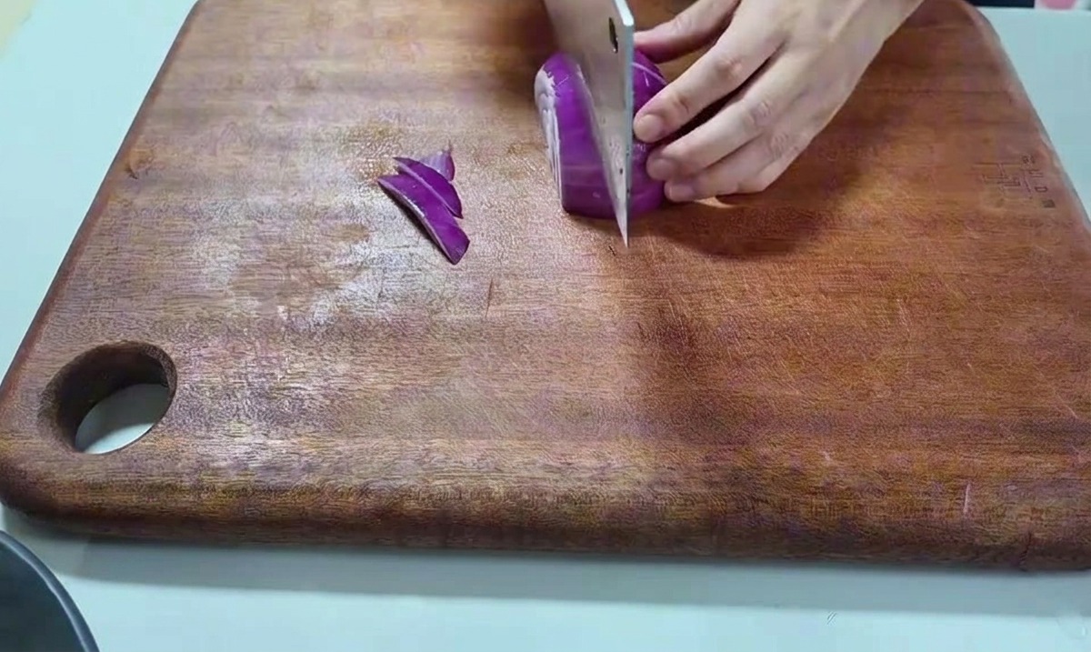 Una mano sujetando una cebolla roja y cortándola con un cuchillo grande sobre una tabla de madera.