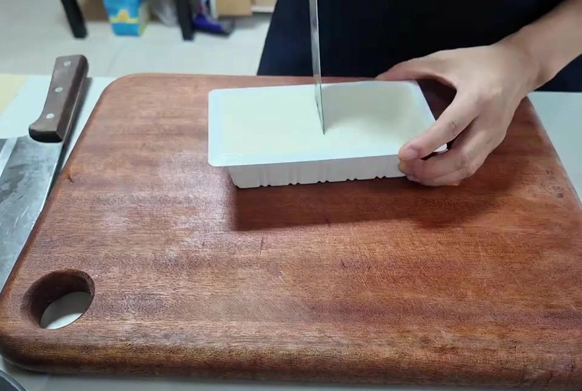 Persona cortando un bloque rectangular de tofu blanco en cubos pequeños sobre una tabla de madera.