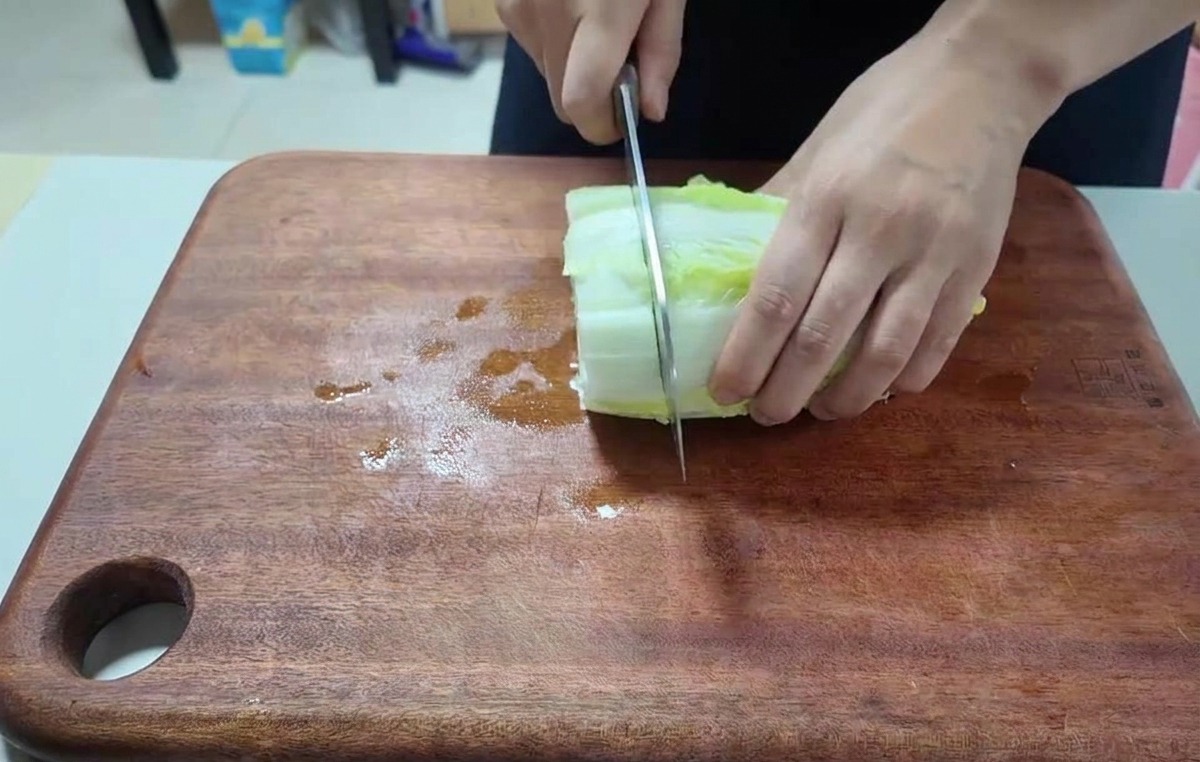 Primer plano de manos usando un cuchillo de chef para picar hojas de col china sobre una tabla oscura.
