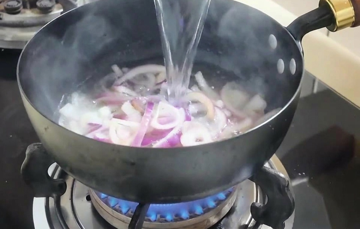 Chorro de agua clara vertiéndose desde un cuenco blanco hacia una sartén con cebollas cocinadas.