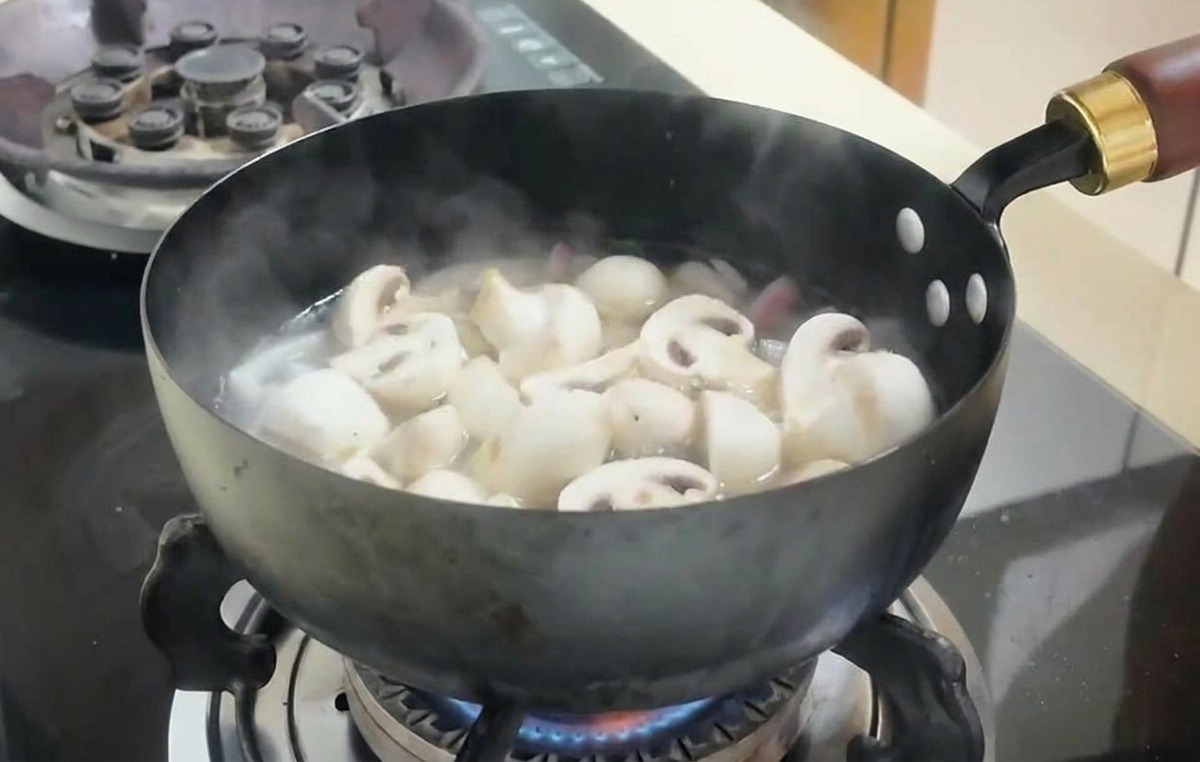 Láminas de champiñones y cubos de tofu hirviendo en un caldo burbujeante dentro de una sartén oscura.