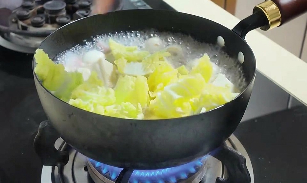 Mano añadiendo granos de maíz amarillo y col picada a una olla de sopa hirviendo.