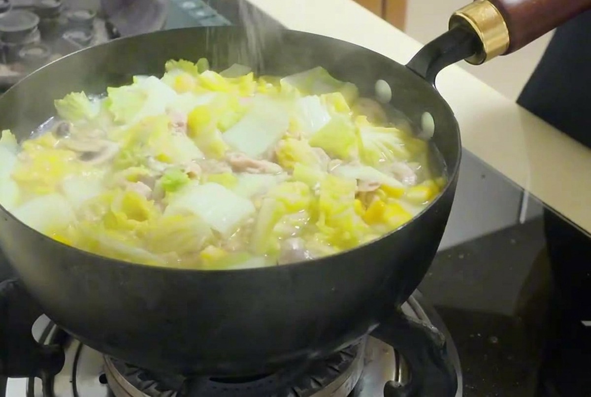 Mano espolvoreando sal fina sobre una sartén humeante con sopa de verduras, tofu y atún.