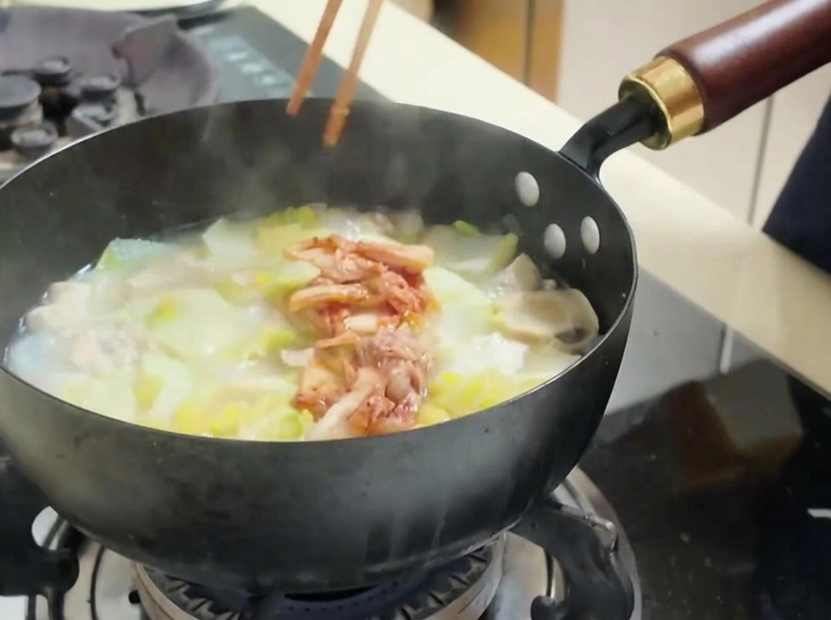 Persona usando palillos para añadir kimchi rojo troceado a una olla de sopa con tofu y col.
