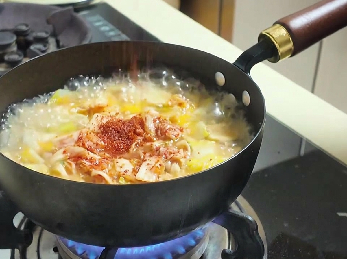 Chile en polvo rojo siendo espolvoreado en una olla de sopa con kimchi y verduras.