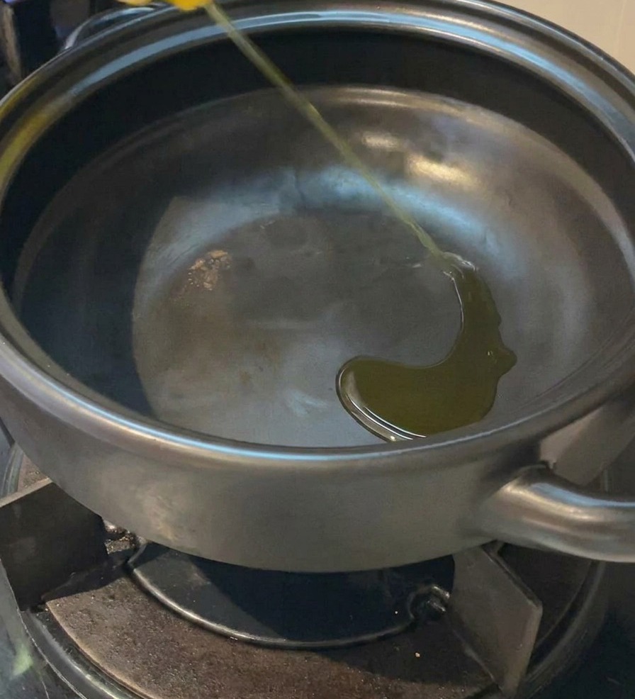 Un fino chorro de aceite vegetal amarillo cayendo al fondo de una olla de barro oscura vacía sobre una estufa de gas.