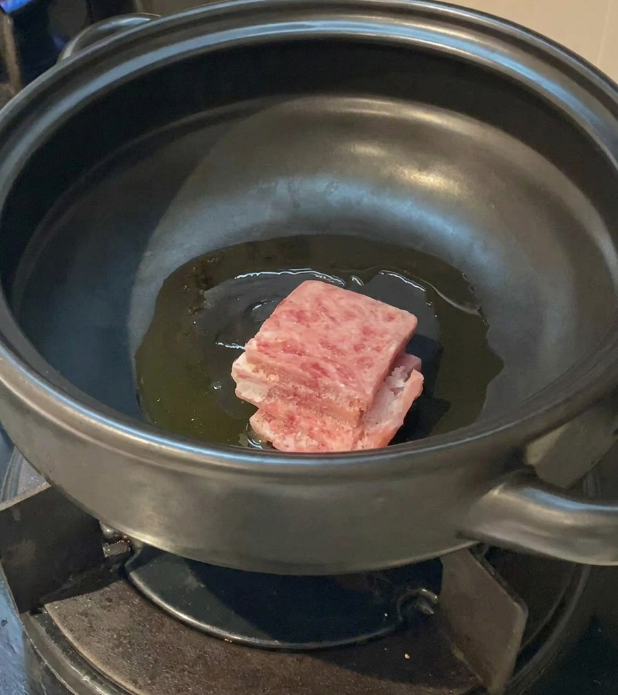 Láminas de panceta de cerdo cruda y marmoleada colocándose en una olla de barro con aceite caliente.