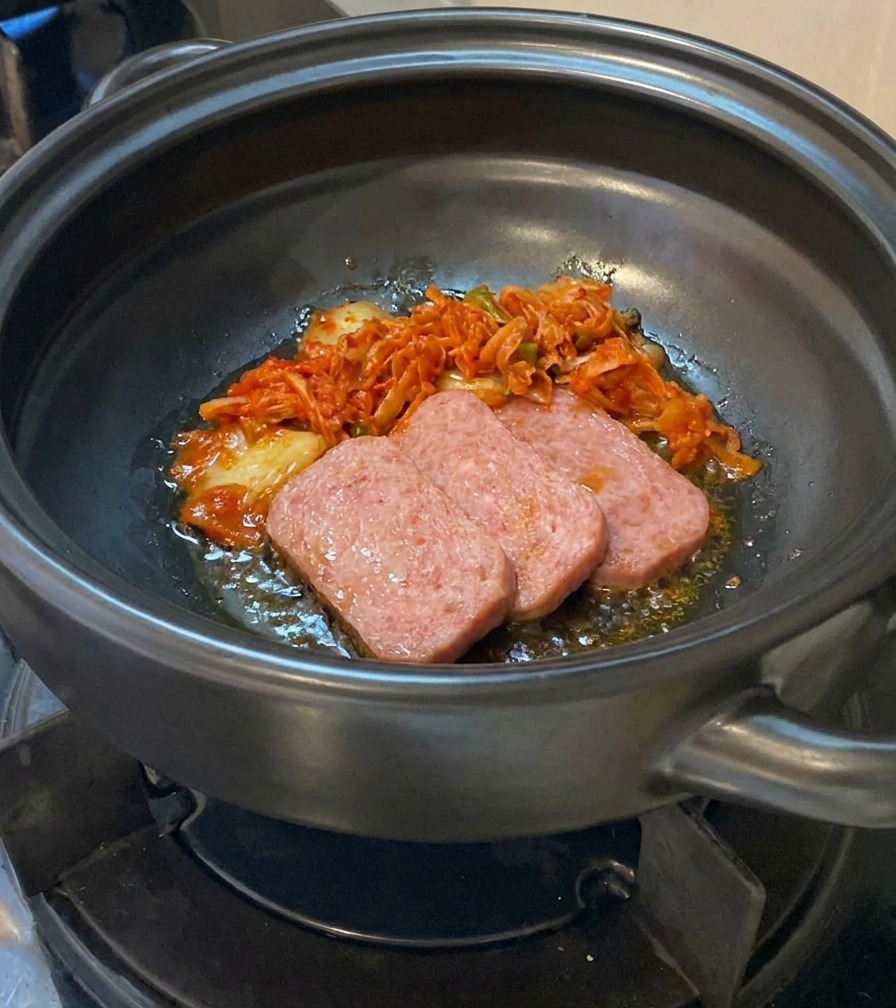 Kimchi picado añadiéndose a una olla de barro que contiene láminas de carne parcialmente cocinadas.