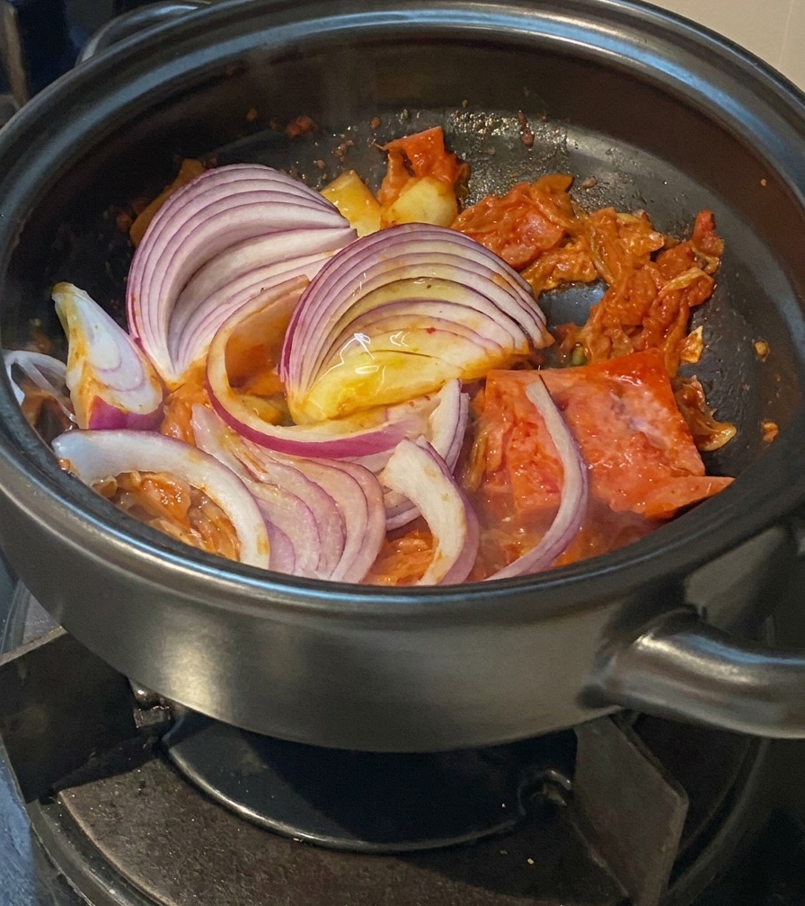 Crecientes gruesas de cebolla roja incorporándose a una olla con kimchi y cerdo salteados.