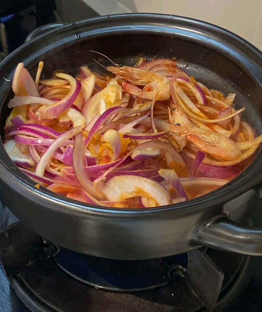 Langostinos frescos enteros con cabeza siendo añadidos a una olla llena de cebolla, kimchi y cerdo.