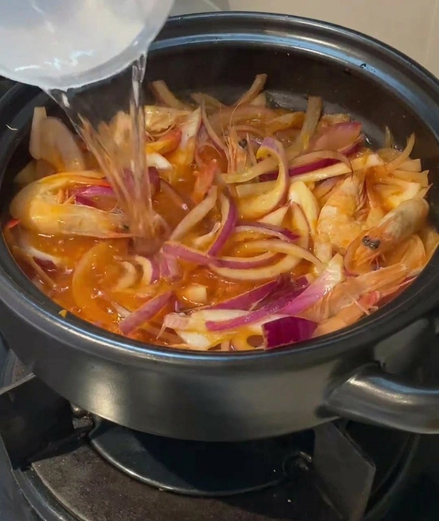 Agua clara vertiéndose desde un recipiente en una olla de barro llena de langostinos, cebolla y kimchi.