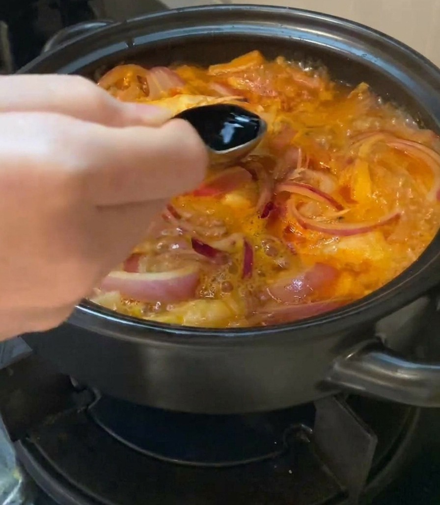 Una mano sosteniendo una cuchara metálica y vertiendo salsa de soja clara en una olla burbujeante de sopa de kimchi.