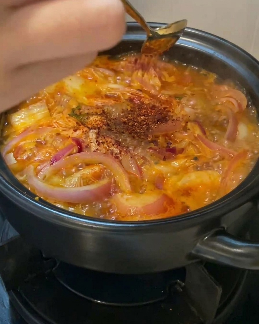 Una mano usando una cuchara dorada para espolvorear chile rojo en polvo sobre una olla burbujeante de sopa de kimchi y cebolla.