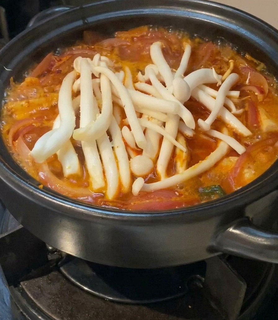 Un grupo de setas frescas blancas descansando sobre la superficie de una sopa de kimchi roja en una olla cerámica negra.