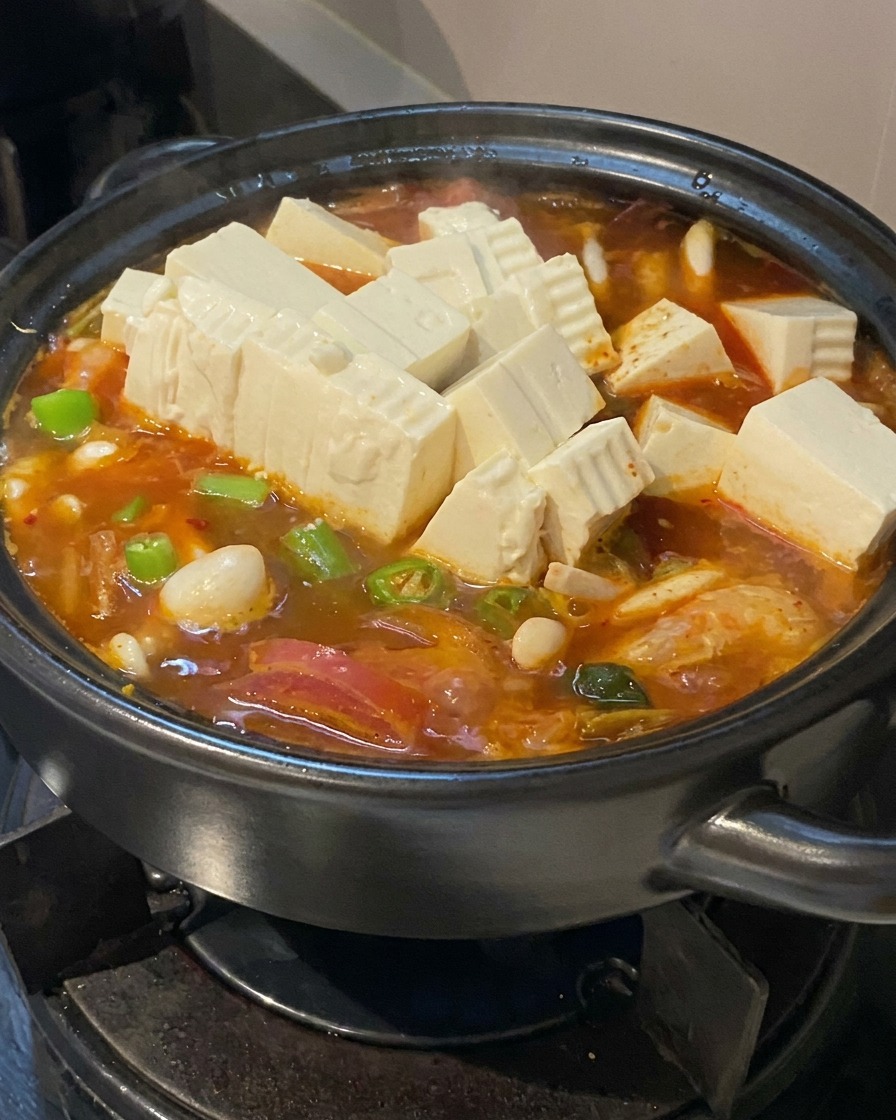 Numerosos cubos de tofu blanco fresco y rodajas de chiles verdes añadiéndose a una sopa burbujeante naranja en una olla negra.