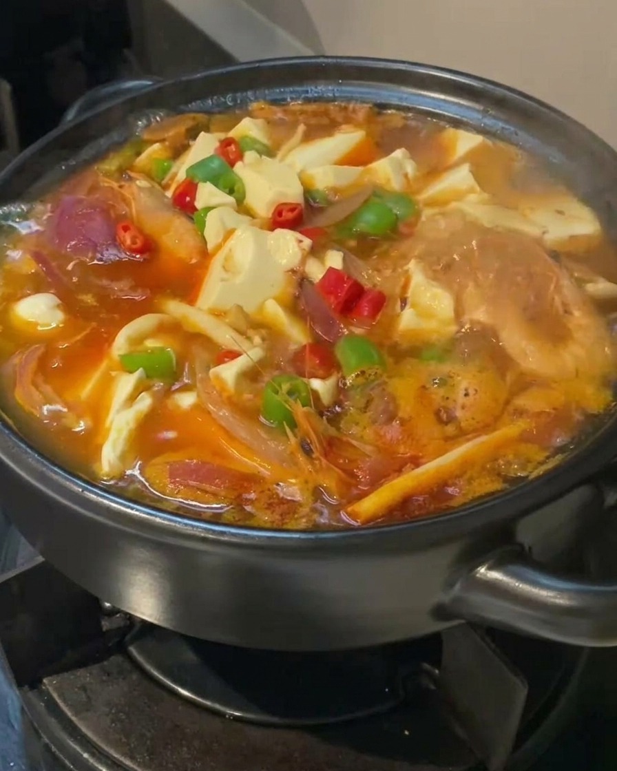 Primer plano de una sopa de tofu y kimchi hirviendo en una olla negra, decorada con chiles rojos y verdes sobre cubos de tofu blanco.