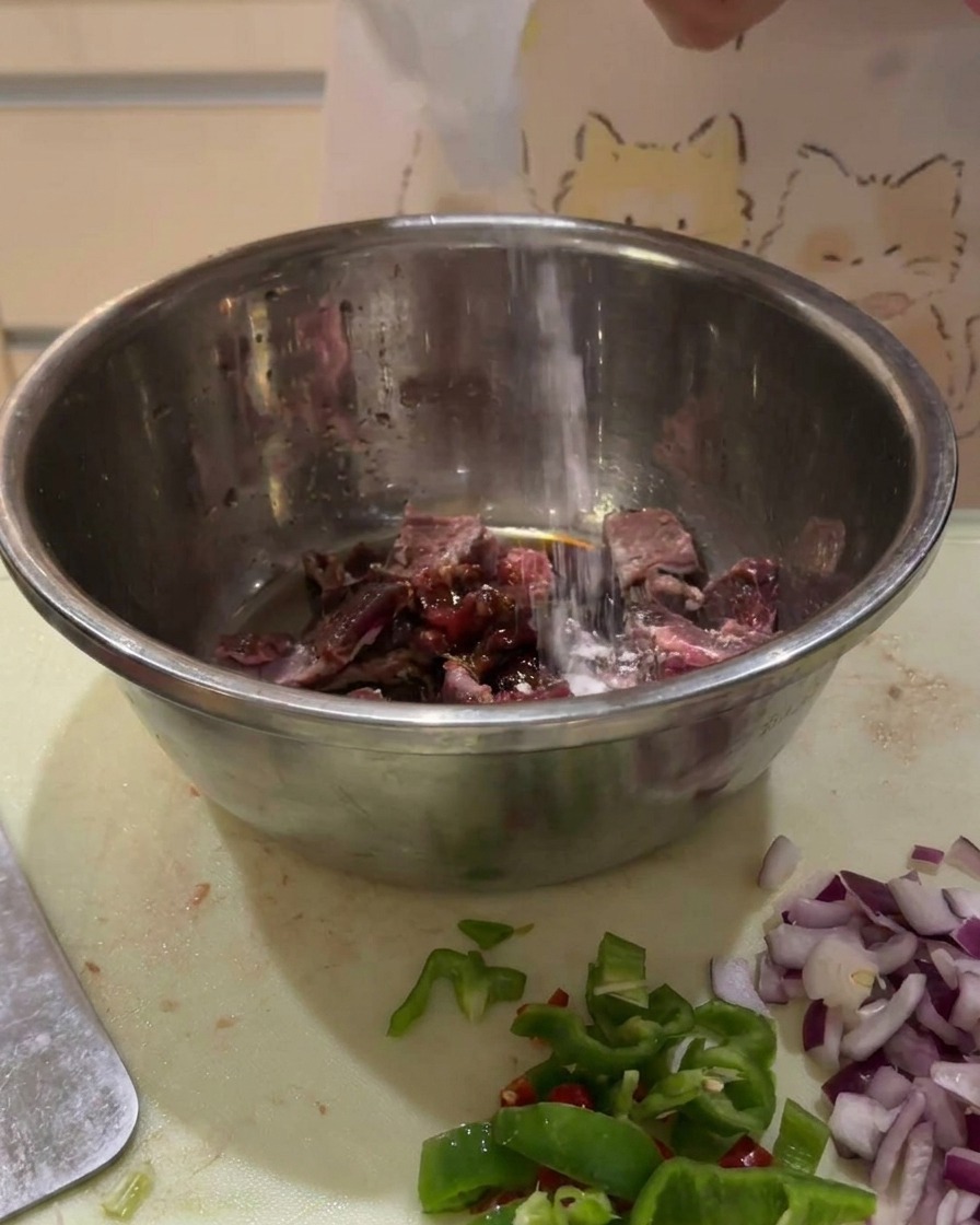 Salsas oscuras siendo vertidas desde una botella sobre cubos de ternera cruda en un bol de metal.