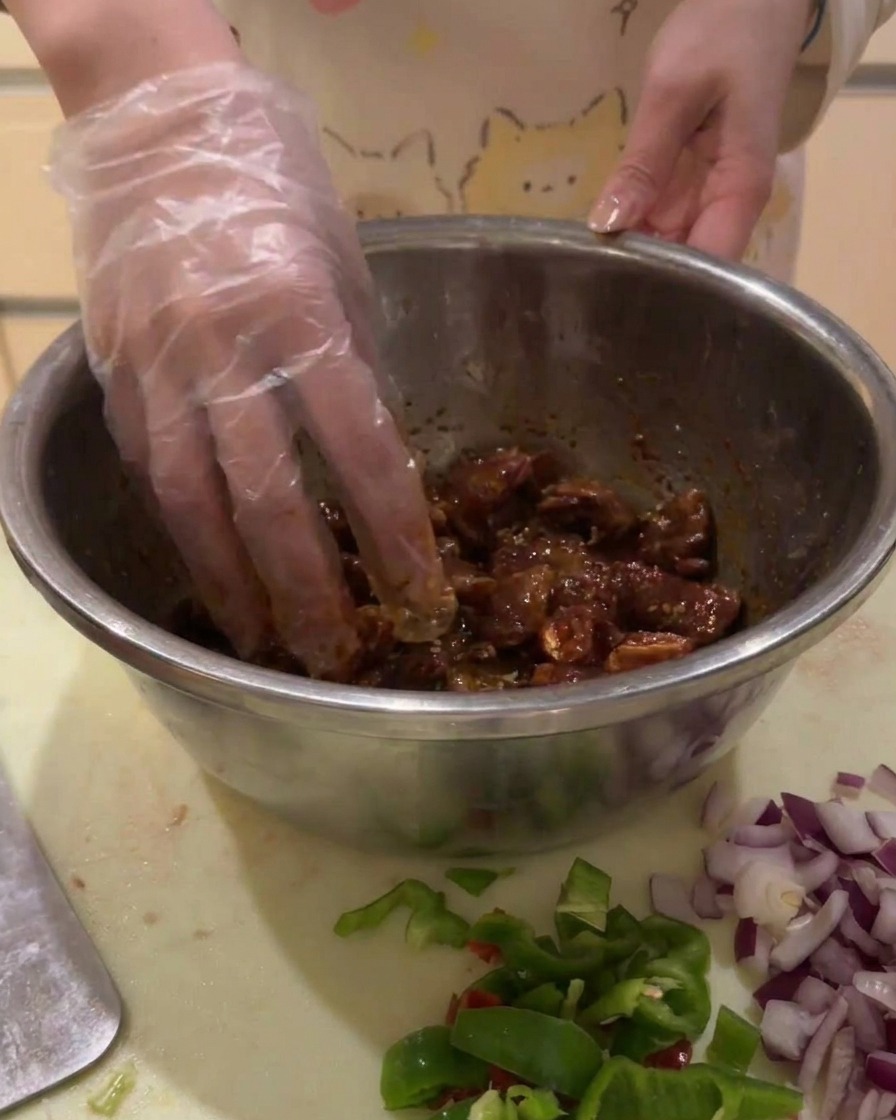 Una mano con guante mezclando los cubos de ternera marinada en un bol plateado para asegurar que se cubran uniformemente.