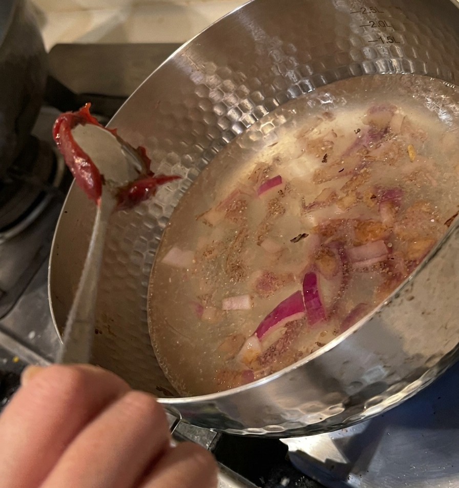 Una cuchara de metal sumergiendo una porción de espesa pasta de chile rojo en la olla con agua y cebolla.