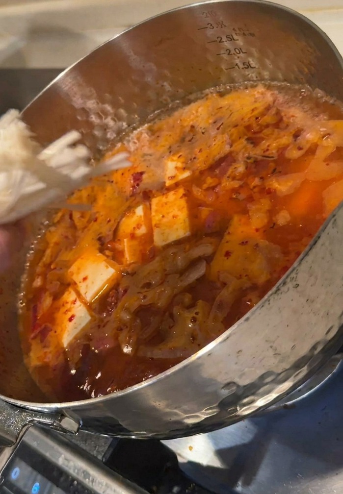 Una mano añadiendo un racimo de setas enoki blancas a una olla burbujeante de sopa roja de kimchi con dados de tofu.