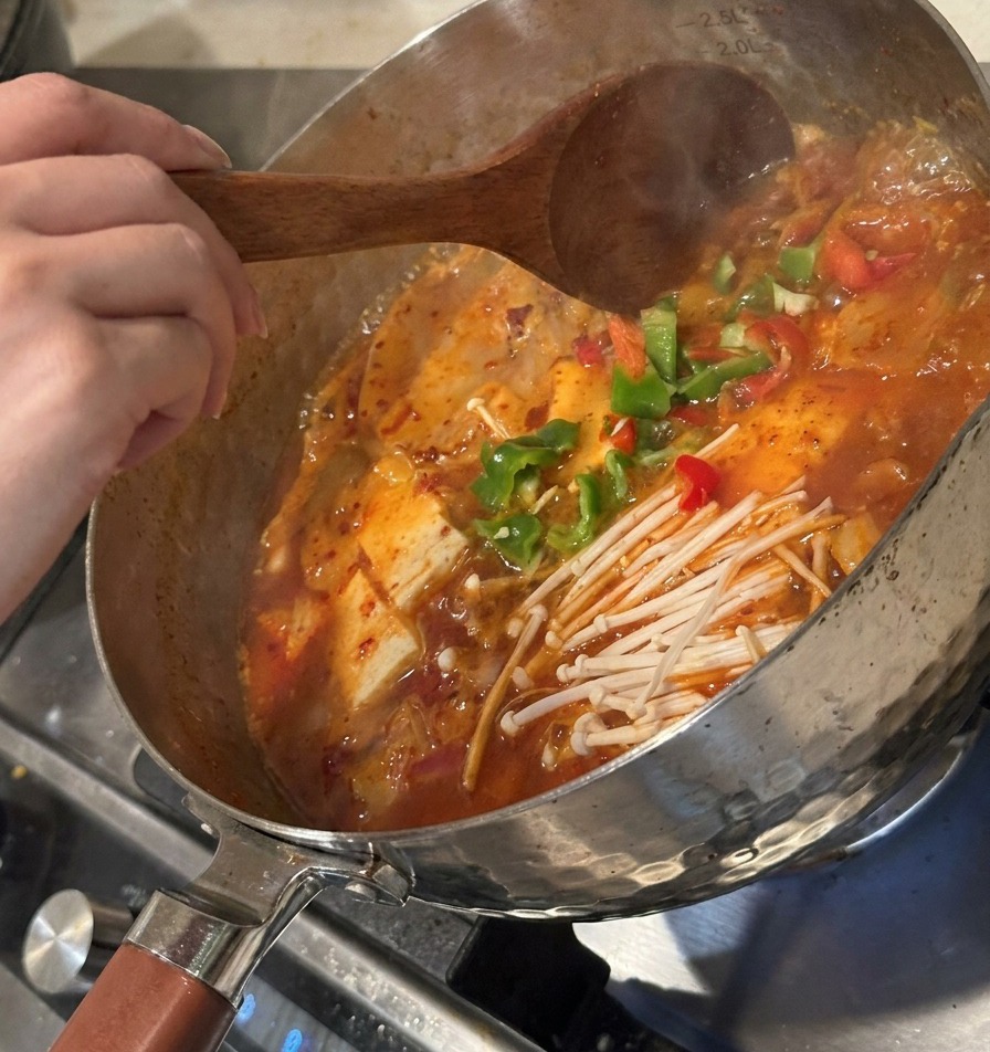 Pimientos verdes en rodajas y kimchi fermentado añadiéndose a la sopa burbujeante y removiéndose con una cuchara de madera.