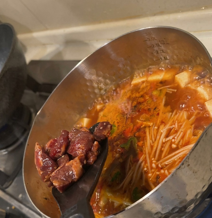 Cubos de ternera marinada cruda empujándose desde una espátula negra a una sopa roja hirviendo llena de verduras.