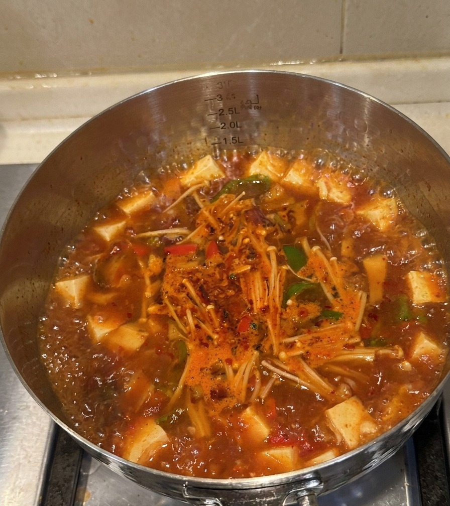Una olla de acero inoxidable llena de sopa de ternera y kimchi burbujeante, mostrando carne cocida, setas enoki y pimientos.