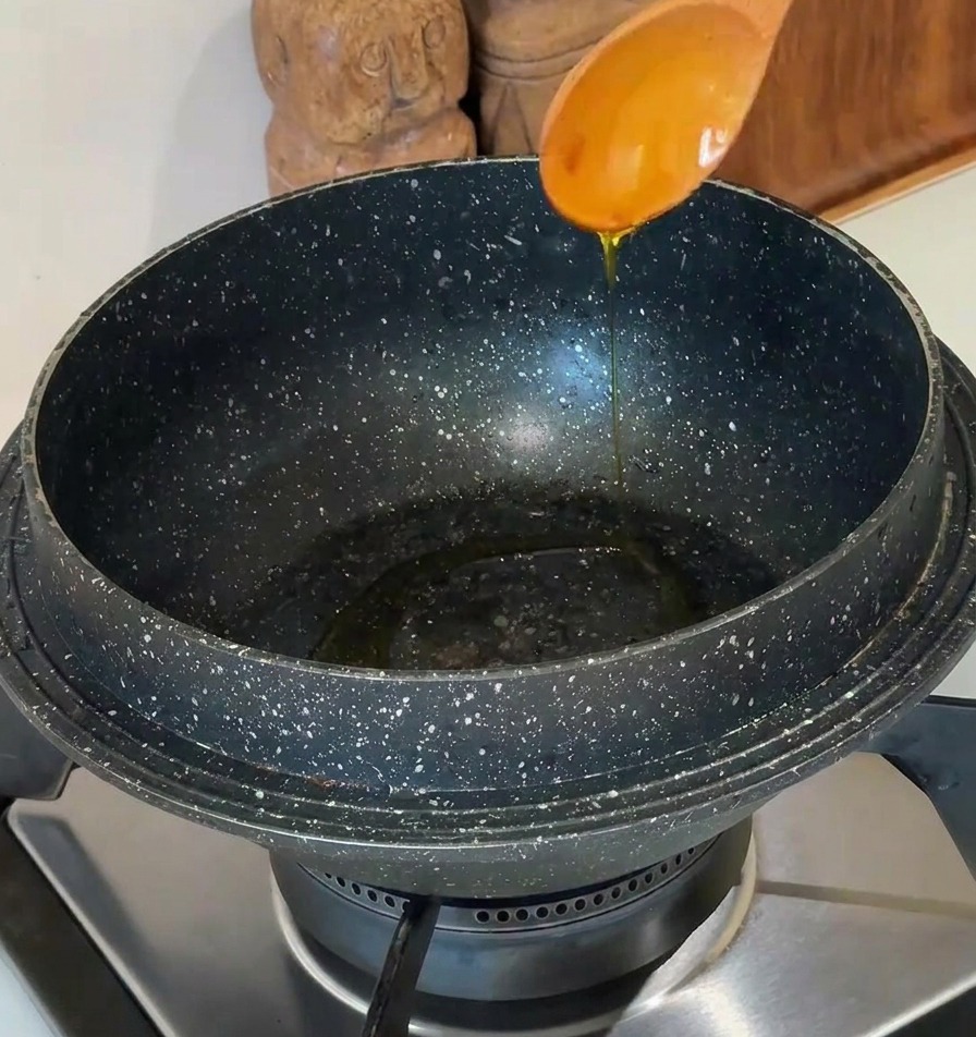 Aceite de cocina vertiéndose desde una cuchara de madera clara en una olla de piedra oscura sobre una estufa moderna.