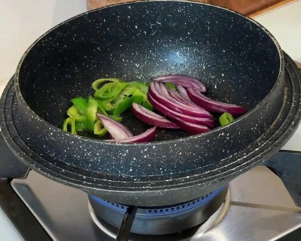 Rodajas de cebolla roja añadiéndose a las cebolletas salteadas en una olla negra sobre una llama de gas azul.